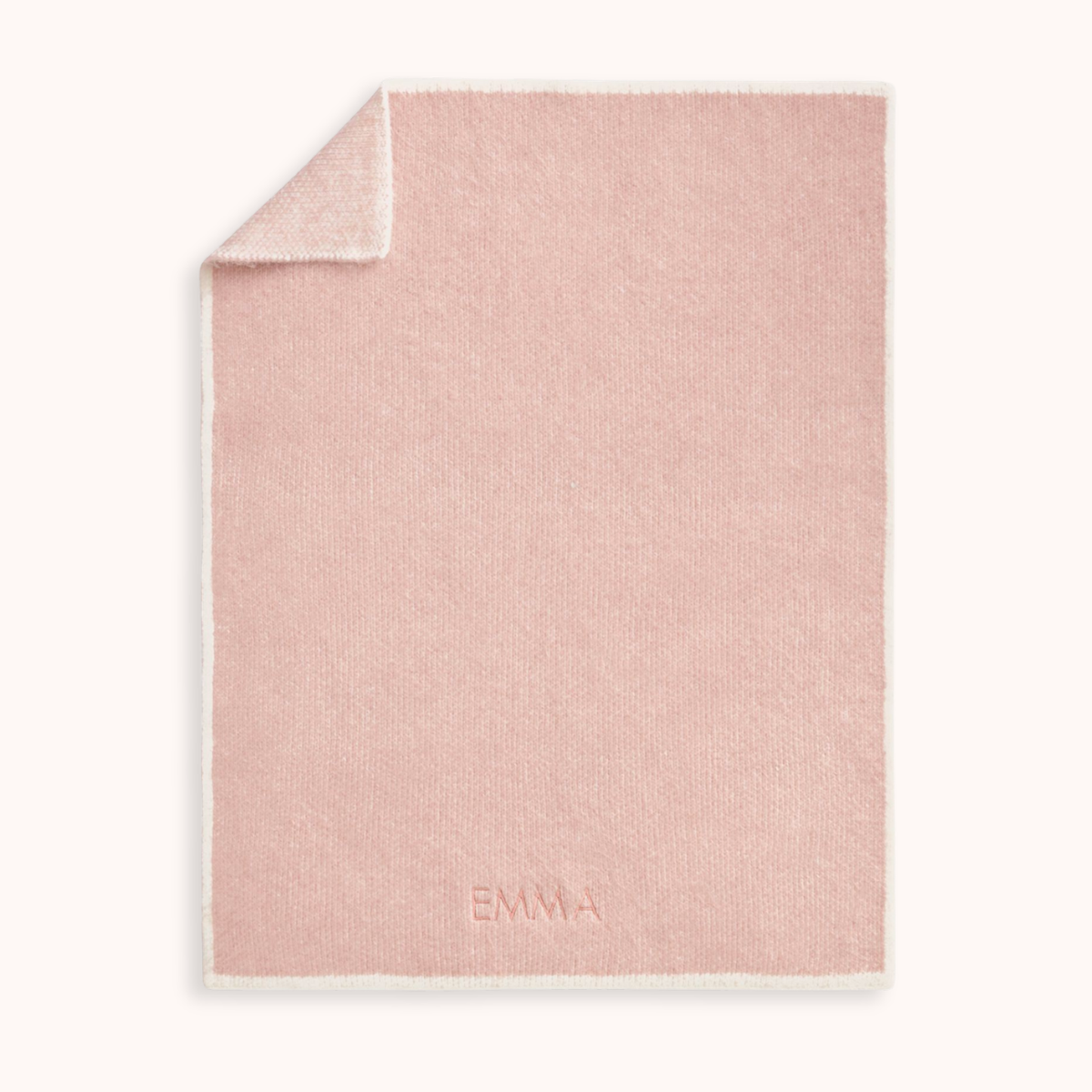 A dusty pink knitted blanket with a white border, featuring the name 'EMMA' embroidered in a matching pink tone near the bottom. The top-left corner is folded over to show the reverse side, set against a clean off-white background.