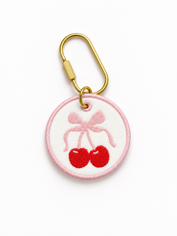 A circular embroidered patch featuring two red cherries tied with a pink bow, attached to a gold carabiner clip on a white background.
