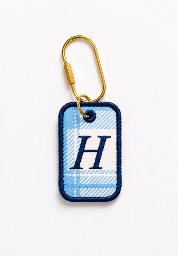 A custom rectangular embroidered key tag with a light blue and white plaid pattern, featuring a navy blue border and a large embroidered letter 'H' in navy. It is attached to a gold metal carabiner clip and photographed on a clean white background.