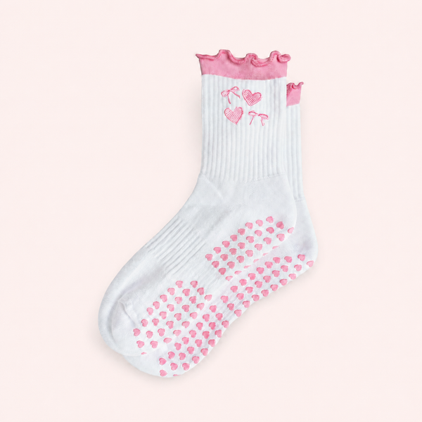 A single white grip sock with pink heart-patterned non-slip dots on the sole, a pink ruffled trim at the top, and pink embroidered cherub motifs, shown on a pale pink background.