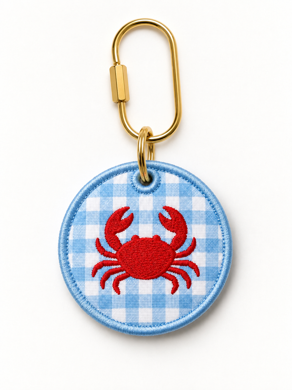A custom circular embroidered patch featuring a red crab on a light blue and white gingham background, attached to a gold metal carabiner clip, shown on a clean white background.