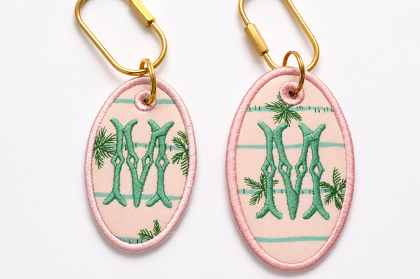 Two oval custom embroidered keychains with gold key rings, featuring a pink and mint green striped background, green monogram letter 'M' embroidery, and pine branch accents, shown on a white background.