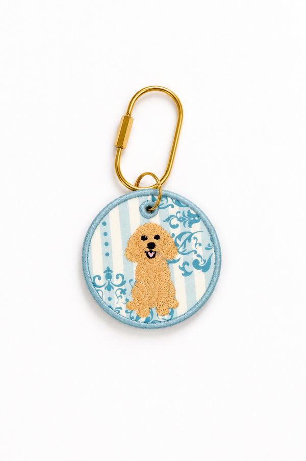 A custom round embroidered dog keychain tag featuring a golden poodle on a light blue and white striped background with blue scrollwork, attached to a gold metal carabiner on a white background.
