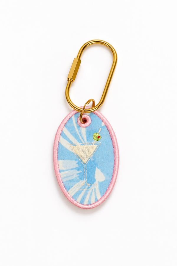 An oval embroidered patch keychain featuring a light blue background with a white sunburst pattern, a pink stitched border, and an embroidered martini glass with a green olive. The patch is attached to a gold carabiner on a clean white background.