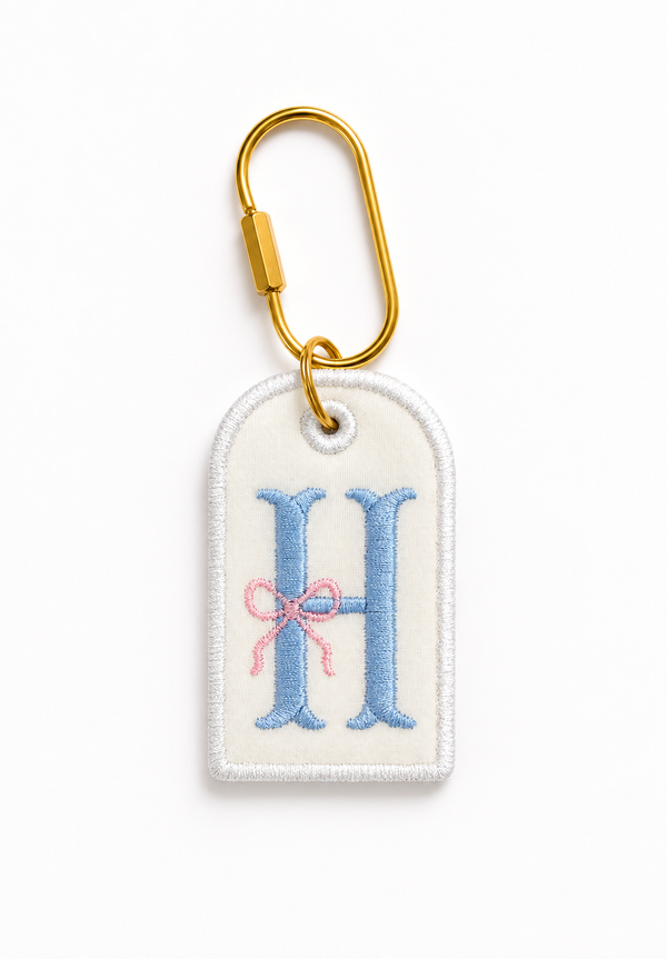 A customized embroidered personalized gift tag keychain featuring a white fabric tag with a large light blue letter 'H' and a small pink bow, attached to a gold metal carabiner on a clean white background.