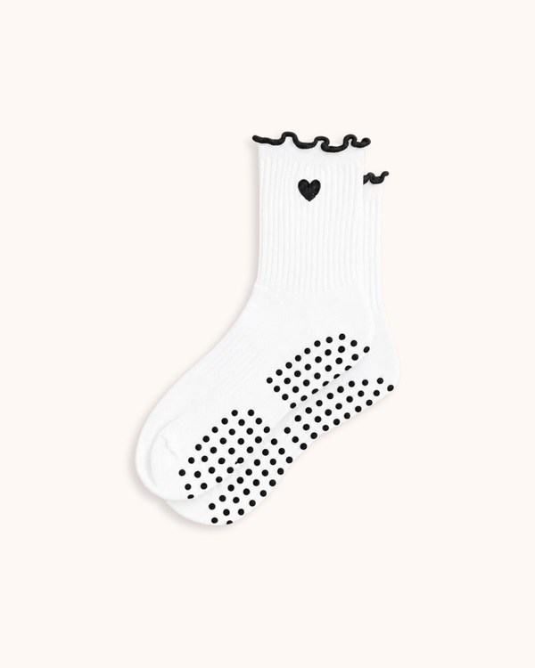 A single white ribbed sock with black grip dots on the sole, a small black heart, and black wavy trim at the top, shown on a plain light beige background.