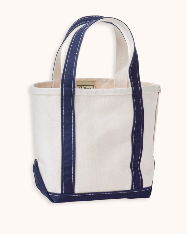 A canvas tote bag with a white body and navy blue trim, featuring thick white contrast stitching and dual top handles, shown against a plain off-white background.