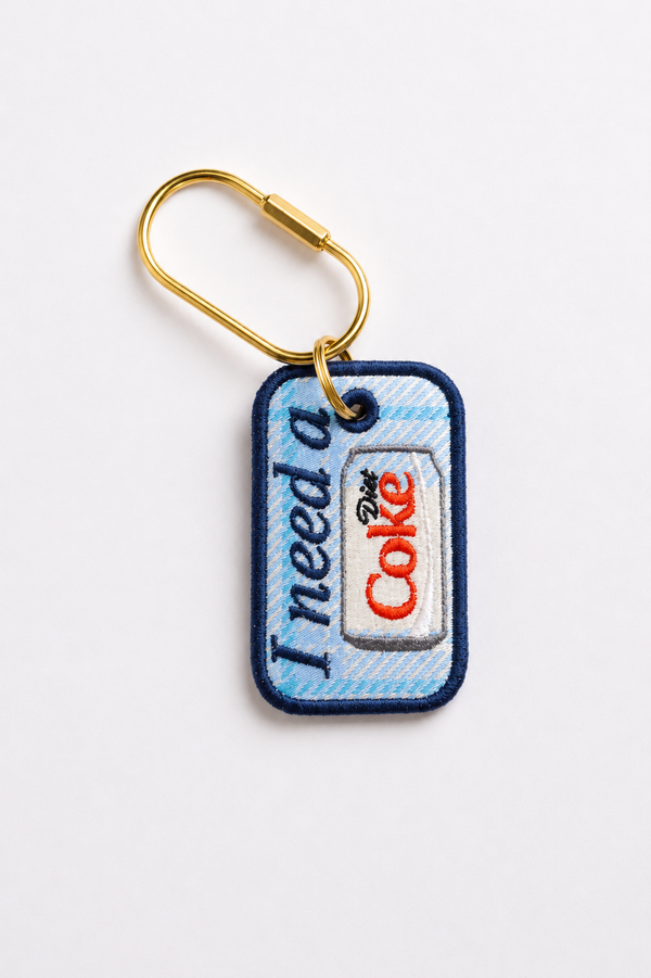 A rectangular custom embroidered keychain tag with a gold split ring and lobster clasp. The tag features a light blue and white diagonal patterned background with navy blue stitched borders.