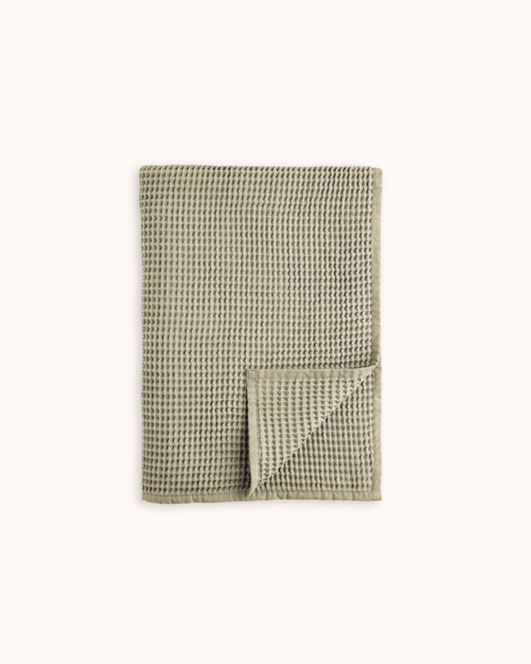 A folded waffle-weave fabric item, likely a tea towel or napkin, in a muted sage green color, displayed on a clean off-white background.