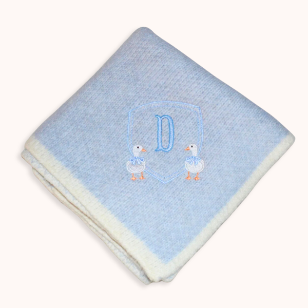 A folded light blue baby blanket with a cream sherpa border, featuring an embroidered blue monogram 'D' inside a shield outline with two white ducks wearing blue bows.
