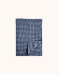 A folded blue waffle-weave cloth napkin or tea towel with a small matching square folded over the corner, displayed on a clean off-white background.