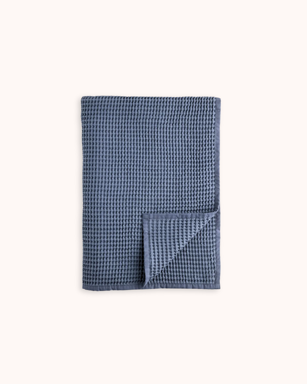 A folded blue waffle-weave cloth napkin or tea towel with a small matching square folded over the corner, displayed on a clean off-white background.