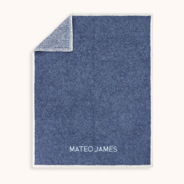 A personalized blue knit blanket with a white border and the name 'MATEO JAMES' embroidered in white at the bottom, shown on a light cream background with one corner folded over.