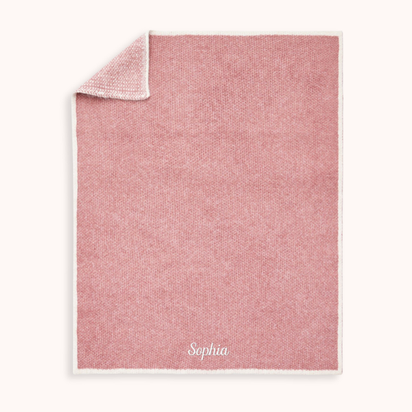 A personalized dusty pink woven blanket with a white border and the name 'Sophia' embroidered in white at the bottom, shown on a light cream background.