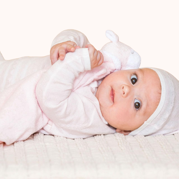 A baby lying on a textured cream blanket, wearing a light pink long-sleeve swaddle and a matching white knit bonnet.