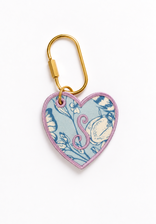 A customized heart-shaped keychain featuring a light blue and pink floral pattern with a pink monogram letter 'S', attached to a gold metal carabiner on a white background.